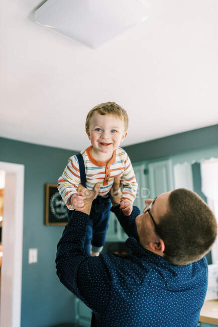 Père soulevant son bébé garçon riant debout dans sa cuisine — Photo de stock