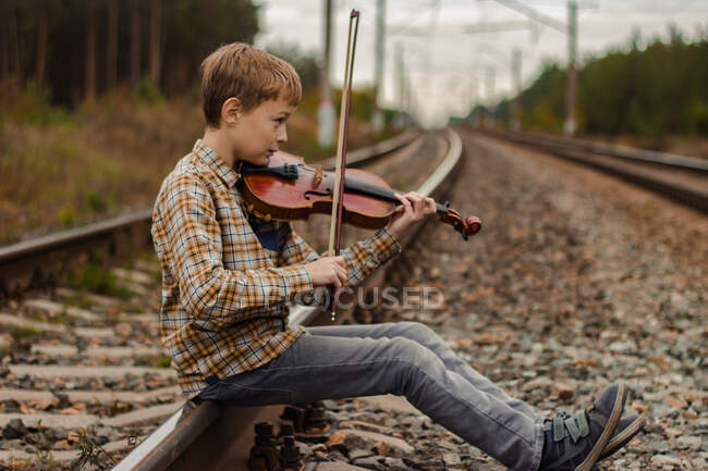 Un hermoso niño rubio se sienta en los rieles del ferrocarril y toca el v - foto de stock
