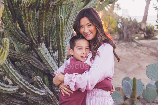 Madre abrazando al hijo delante de un gran cactus, ambos mirando a la cámara. - foto de stock