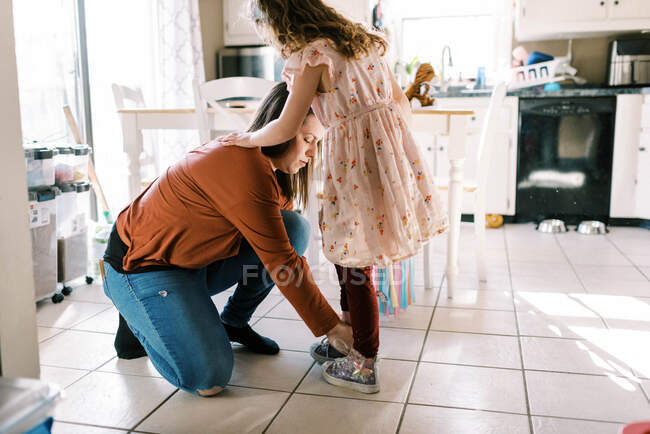 Mother helping her daughter put on shoes with laces in kitchen — Stock Photo