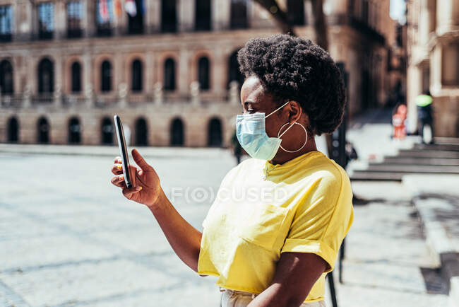 Retrato de menina negra com máscara facial, cabelo afro e brincos de argola usando seu celular em uma praça velha. — Fotografia de Stock