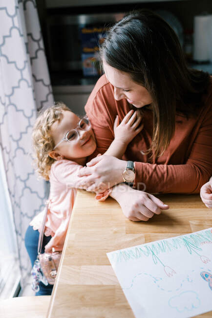 Una bambina con gli occhiali che abbraccia sua madre al tavolo della cucina — Foto stock