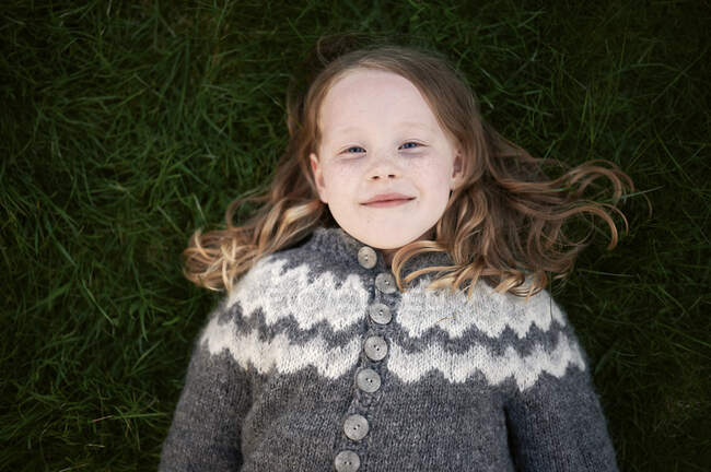 Visão superior da menina pálida em jaqueta de malha deitada no gramado gramado e olhando para a câmera com sorriso — Fotografia de Stock