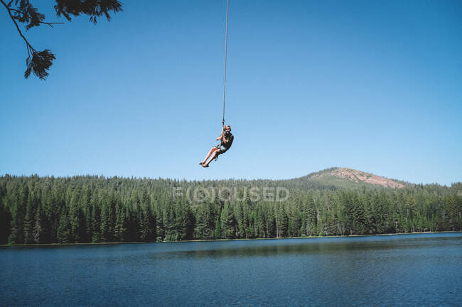 Chico joven se balancea por encima del lago en un columpio de cuerda - foto de stock