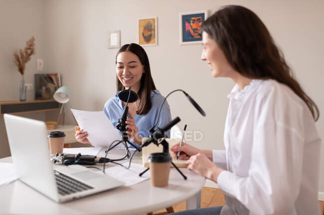 Happy multiethnic women reading and making notes for podcast together in studio — Stock Photo