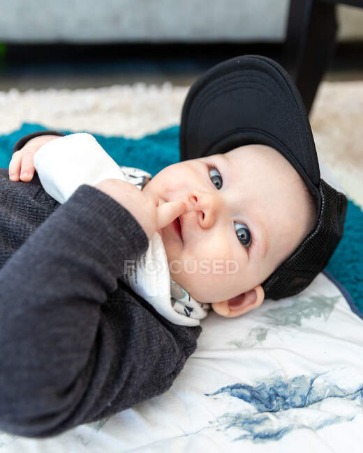 Curious baby boy laying down wearing baseball cap. — Stock Photo