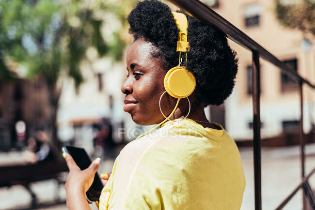Вид сзади на чёрную девушку с афроволосами и серьгами, слушающую музыку с помощью мобильного телефона и жёлтых наушников в городском пространстве в городе. — стоковое фото