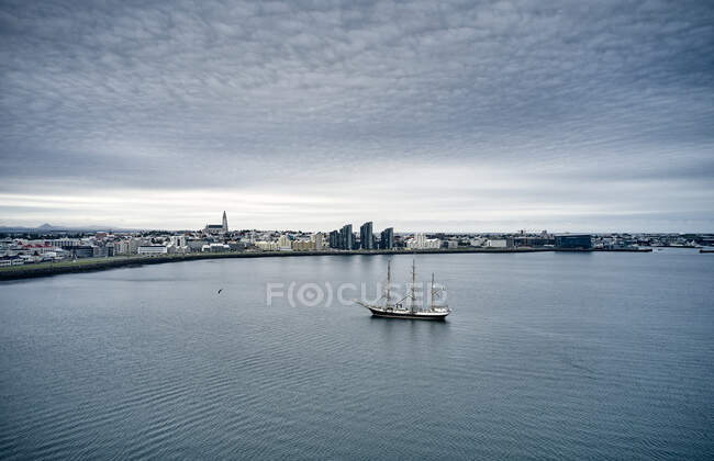 Вид на корабель з щоглами, що пливуть по морській воді біля прибережної зони міста проти похмурого неба в похмурий день — стокове фото