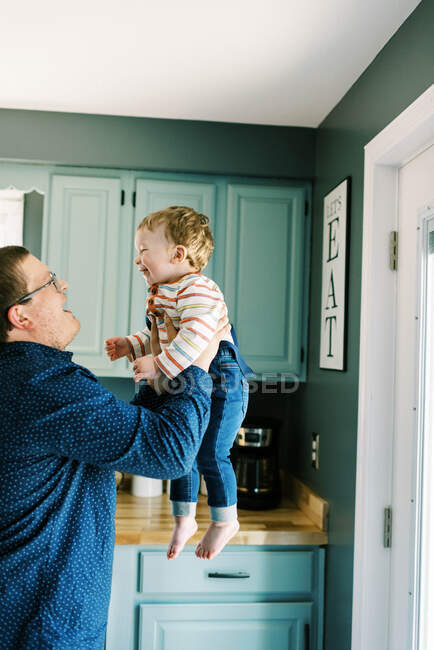 Pai segurando seu menino no ar de pé em sua cozinha — Fotografia de Stock