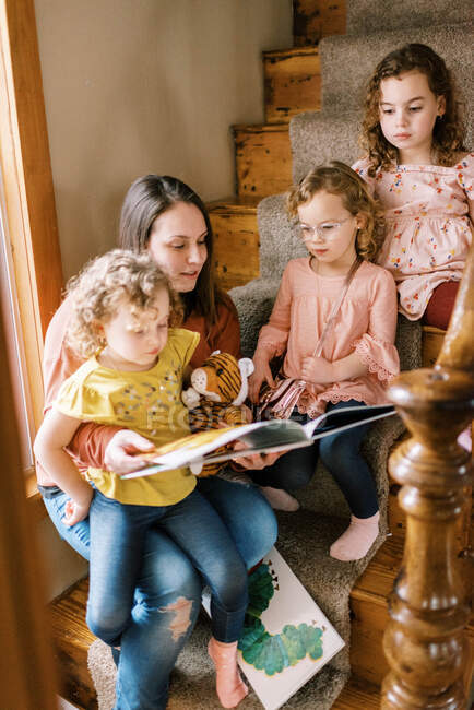 Una joven madre y sus tres hijas sentadas en la escalera leyendo libros - foto de stock
