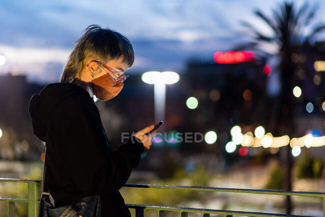 Adolescente com óculos olhando para o telefone usando máscara no cenário da cidade — Fotografia de Stock