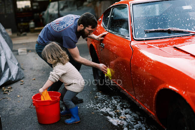 Niña limpiando un coche clásico rojo con su padre juntos - foto de stock