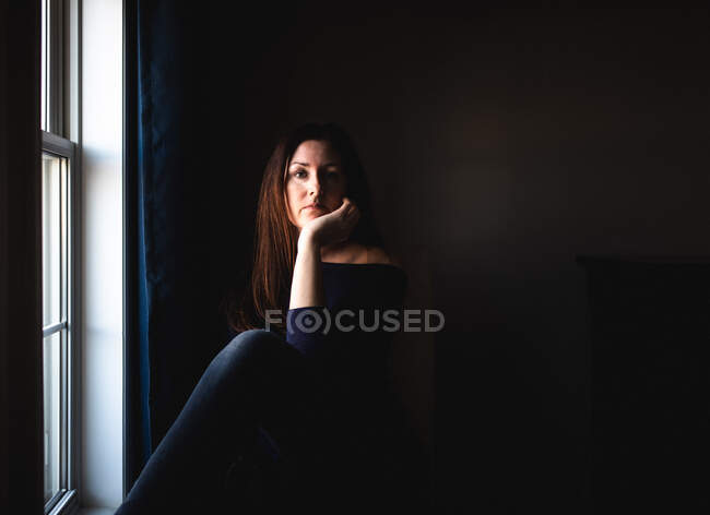 Привлекательная женщина, сидящая одна в темной комнате у окна. — стоковое фото