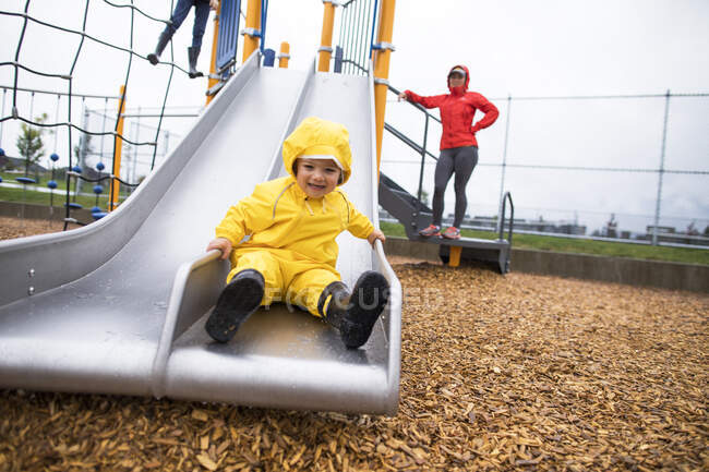 Rapaz feliz deslizando no slide no parque local, mãe assistindo. — Fotografia de Stock