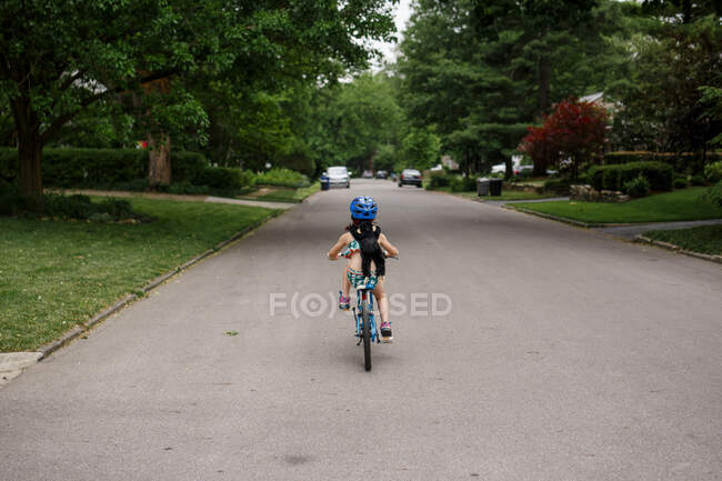 Маленькая девочка едет вниз по улице на велосипеде одна с игрушечной обезьянкой на спине — стоковое фото