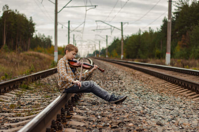 Ein schöner blonder Junge sitzt auf den Schienen der Eisenbahn und spielt Geige. — Stockfoto
