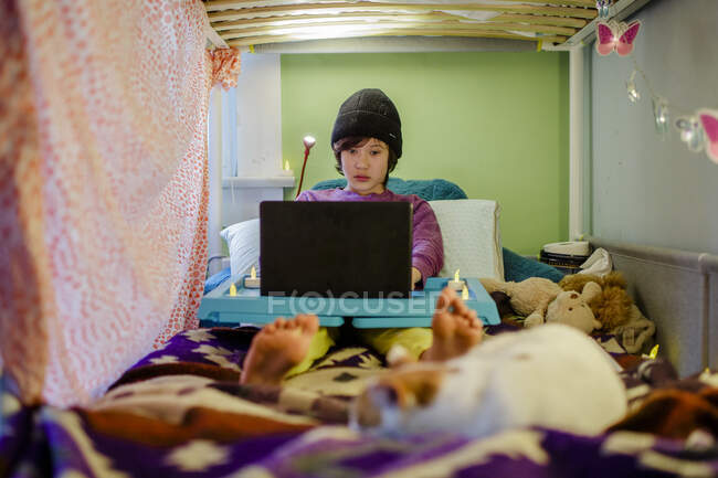 Un ragazzo si siede a letto con il cappello a maglia facendo i compiti sul computer con il gatto — Foto stock