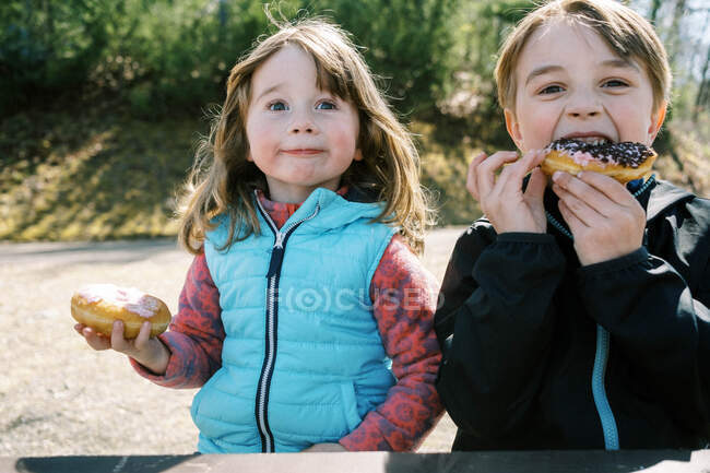 Dos niños sentados en un banco de picnic comiendo rosquillas esmeriladas de fresa - foto de stock