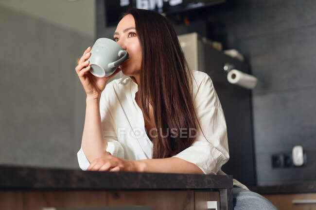 Beautiful smiling woman sitting at the table in the kitchen. Looks into the camera. — Stock Photo