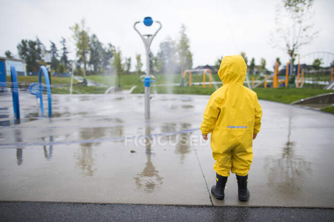 Bambino in tuta antipioggia e stivali guarda il parco acquatico in una giornata umida — Foto stock