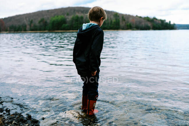 Petit garçon en bottes en caoutchouc rouge marchant à travers l'eau bleue immaculée — Photo de stock