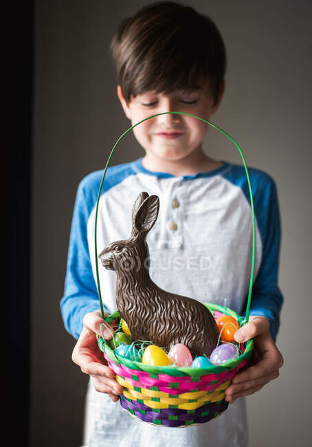 Joven niño feliz sosteniendo cesta de Pascua llena de huevos y conejito de chocolate - foto de stock