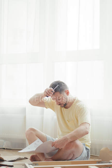 Joven hombre atractivo en una camiseta amarilla ensambla muebles de acuerdo con las instrucciones mientras está sentado en una sala de estar luminosa y aireada. Montaje de muebles en casa. Autoaislamiento, bricolaje. - foto de stock