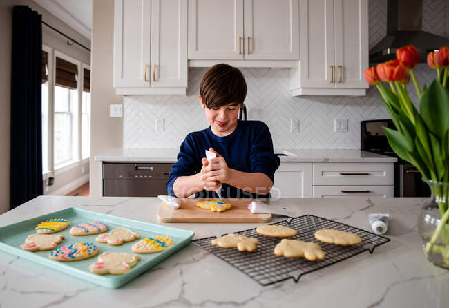 Мальчик украшает пасхальное печенье на прилавке современной кухни. — стоковое фото