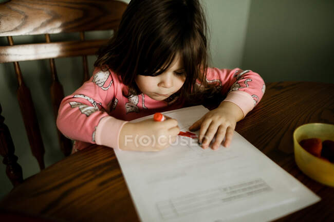 Niña usando pijamas para colorear con un marcador rojo en la mesa de la cocina - foto de stock