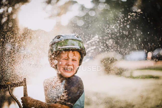 Молодий хлопець грає надворі у спринклері розщеплюючи воду і сміючись — стокове фото