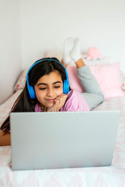 Cheerful teen student using laptop and listening to music on bed — Stock Photo