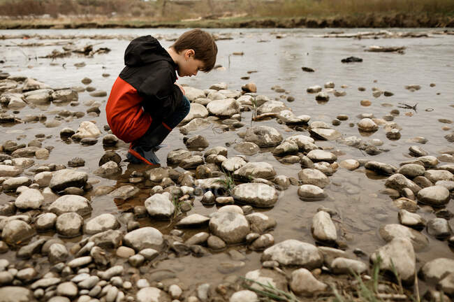 Young boy crouching in a river bed surrounded by rocks in a red jacket — Stock Photo