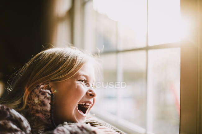 Chica joven con el pelo rubio riéndose delante de la ventana con Sun-Flare - foto de stock