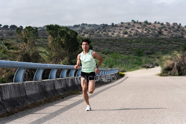 Jovem corredor latino-americano correndo em uma ponte. Estrada e floresta ao fundo. — Fotografia de Stock