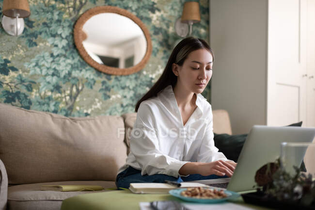 Freelancer asiático jovem navegando netbook enquanto trabalhava em projeto remoto no sofá em casa — Fotografia de Stock