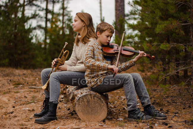 Ragazzo e ragazza, fratello e sorella, gli adolescenti suonano la tromba e il violino seduti nel bosco su un tronco. — Foto stock