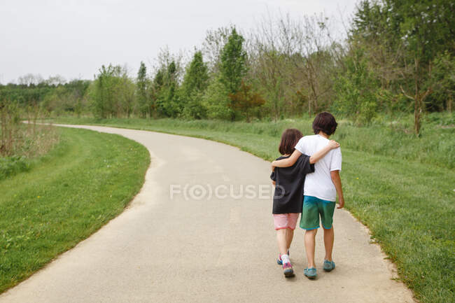 A small boy and girl walk arm in arm down curved path in a park — Stock Photo