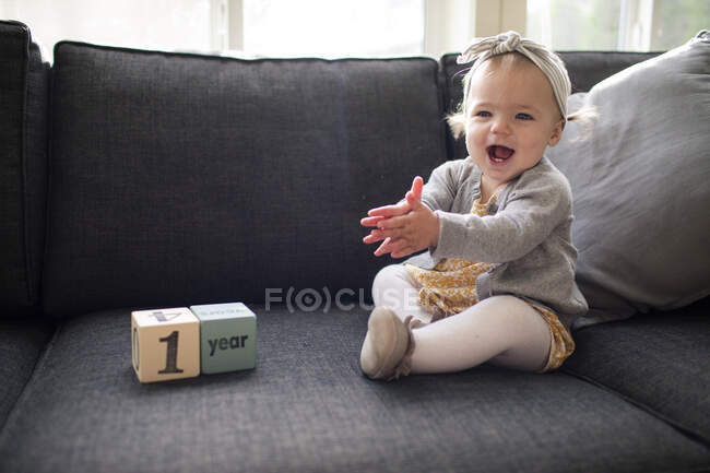 Uma menina de um ano sentada no sofá batendo palmas com emoção — Fotografia de Stock