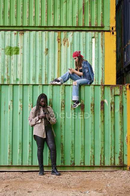 Дві багатонаціональні жінки з телефонами в оточенні морських контейнерів — стокове фото