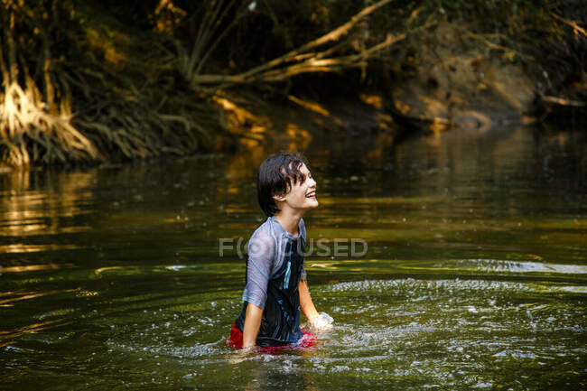 Um menino feliz joga em um rio na luz do sol dourada no verão — Fotografia de Stock