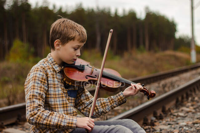 Un hermoso niño rubio se sienta en los rieles del ferrocarril y toca el v - foto de stock
