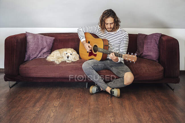 Привлекательный мужчина с длинными волосами играет на акустической гитаре в помещении с собакой — стоковое фото