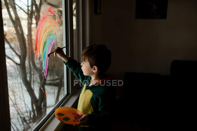 Young boy in costume painting a rainbow on a window in a living room — Stock Photo