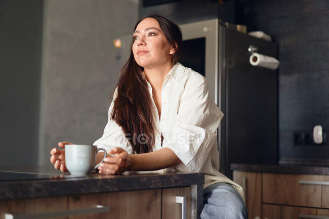 Молода жінка п'є каву з чашки на кухні — стокове фото