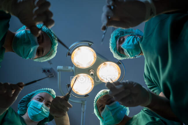 Команда хирургов со скальпелем работают вместе на фоне хирургической лампы, точка зрения снимка. — стоковое фото