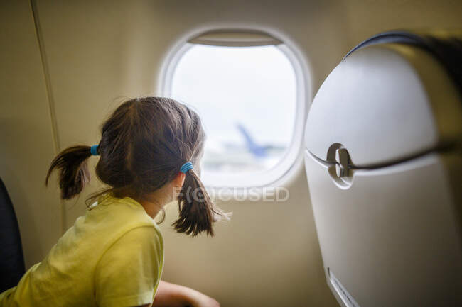 Kleines Mädchen mit Zöpfen sitzt im Flugzeug und starrt aus dem Fenster auf den Asphalt — Stockfoto