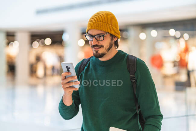 Hombre español moderno sonriendo y usando teléfono inteligente en el centro comercial - foto de stock