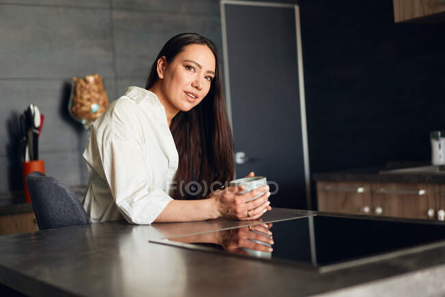 Mujer joven sentada en la cocina con una taza de café - foto de stock