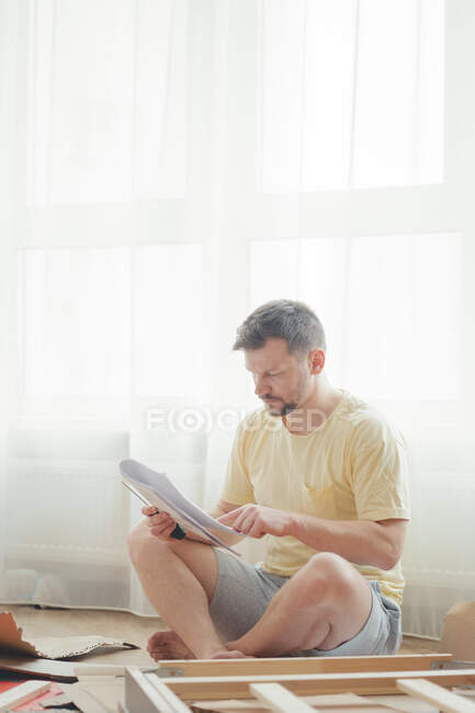 Joven hombre atractivo en una camiseta amarilla ensambla muebles de acuerdo con las instrucciones mientras está sentado en una sala de estar luminosa y aireada. Montaje de muebles en casa. Autoaislamiento, bricolaje. - foto de stock