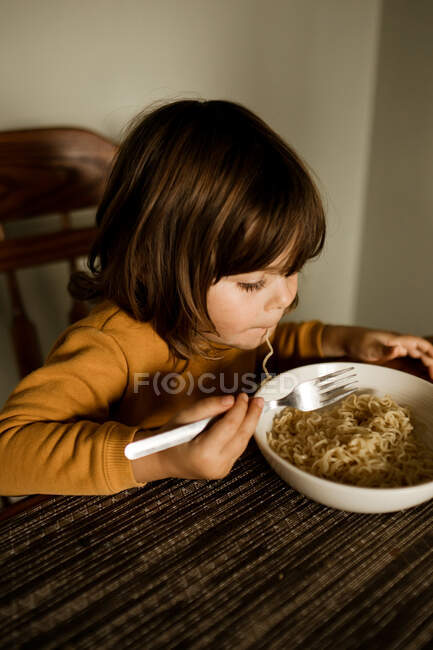 Menina comendo macarrão em uma camisola amarela em sua mesa de cozinha — Fotografia de Stock